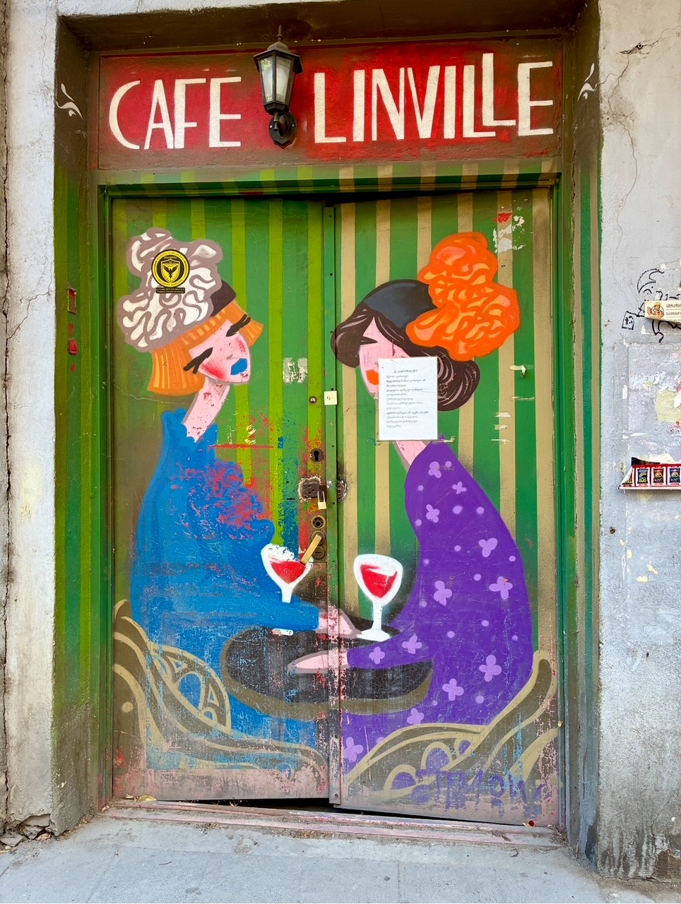 Bemalte grüne Doppeltür mit zwei stilisierten Frauen in blauer und lila Kleidung, die an einem kleinen Tisch mit zwei Weingläsern sitzen, darüber rotes Schild mit weißer Aufschrift 'CAFE LINVILLE'
