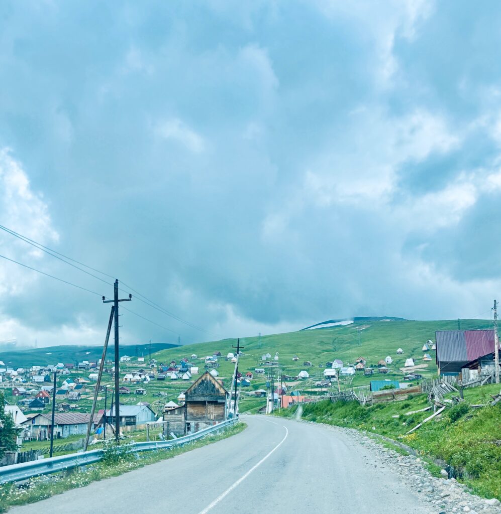 Bergstraße mit Leitplanke führt an einem Dorf mit verstreuten Häusern am Hang unter bewölktem Himmel vorbei