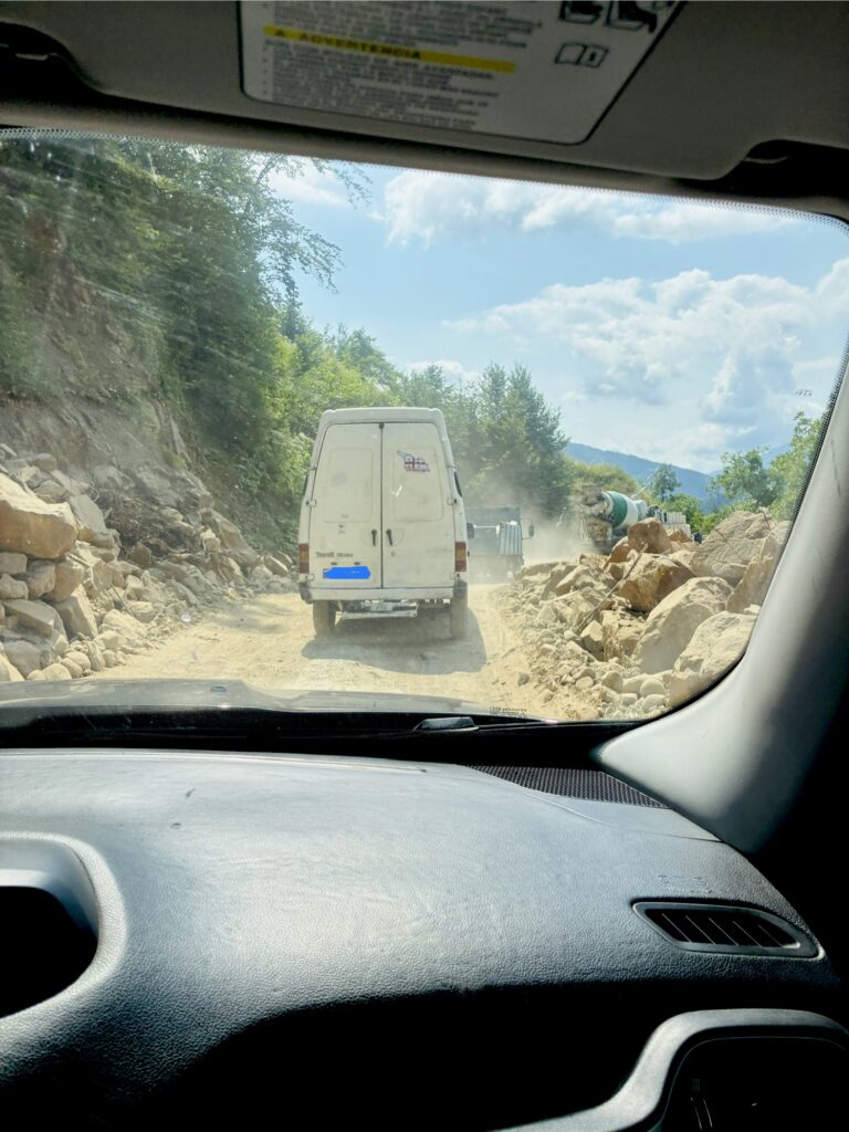 Blick aus einem Fahrzeug auf eine unbefestigte Straße mit einem weißen Transporter und einem Lastwagen vor Staubwolken