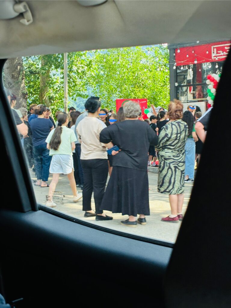 Menschenmenge am Straßenrand vor einem Gebäude mit roten und weißen Luftballons, aufgenommen aus einem Fahrzeug heraus