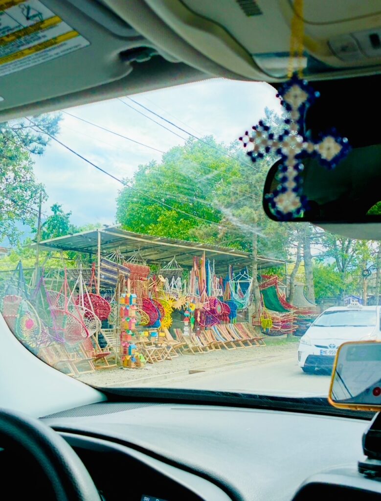 Blick aus einem Auto auf eine Straßenseite mit einem Marktstand, der bunte geflochtene Körbe und Stühle anbietet, im Vordergrund ein hängendes Kreuz am Innenspiegel