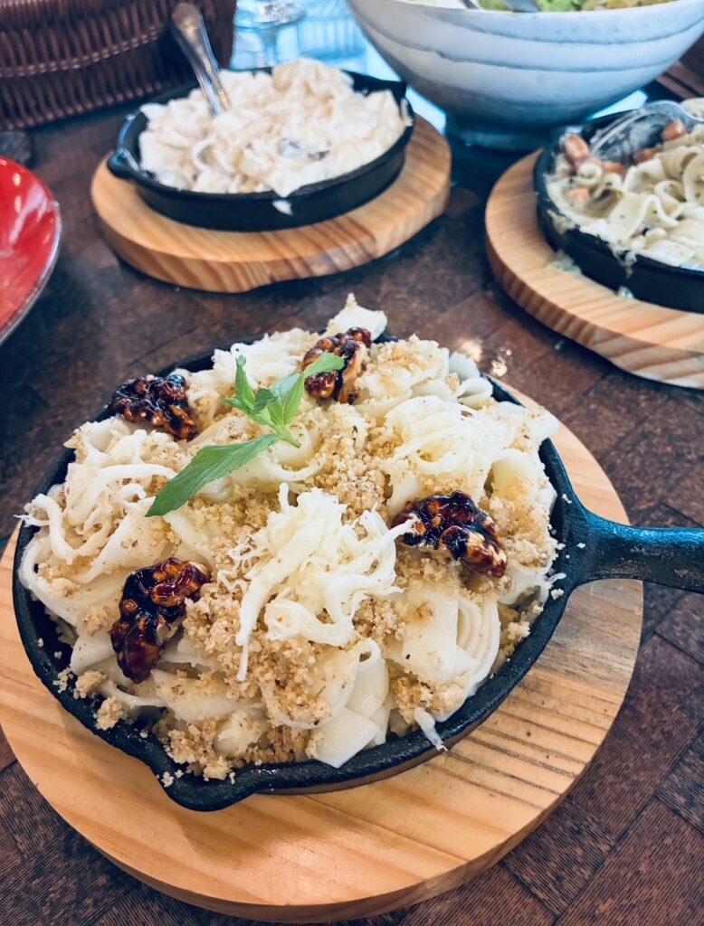 Kleine gusseiserne Pfanne mit Nudelgericht, garniert mit Nüssen und frischem grünen Kraut, auf rundem Holzbrett