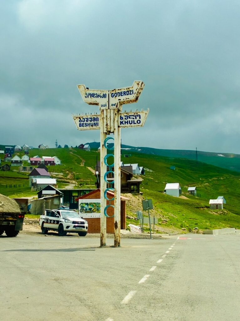 Wegweiser mit vier Schildern in georgischer und lateinischer Schrift auf einer ländlichen Straße mit grünen Hügeln und verstreuten Häusern im Hintergrund