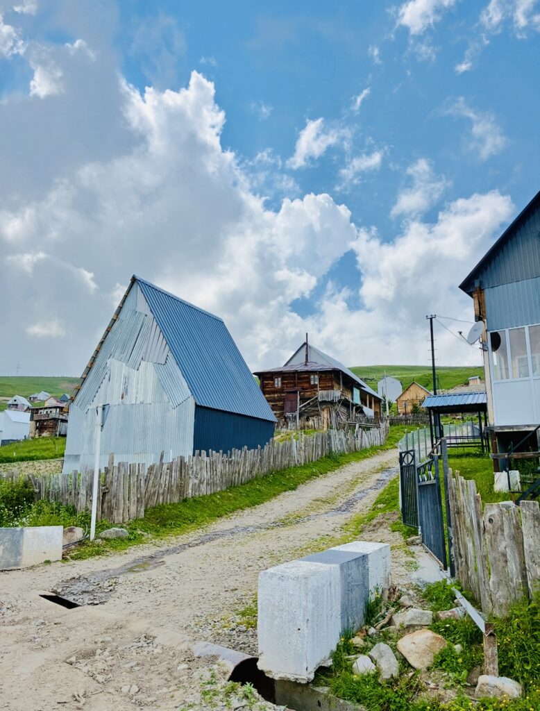 Schräge Wellblechhütte neben einem unbefestigten Weg mit Holzzaun und weiteren Häusern unter blauem Himmel mit Wolken