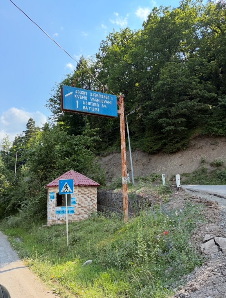 Straßenschild mit Richtungspfeil und den Ortsnamen Kvemo Vashlvani und Batumi in lateinischer und georgischer Schrift neben einem kleinen gemauerten Unterstand an einer kurvigen Straße im Grünen
