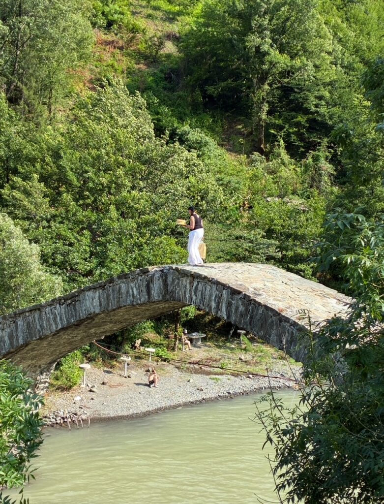 Person in weißer Hose und schwarzem Oberteil steht auf einem steinernen Bogenbrücke über einen Fluss, umgeben von dichtem Grün