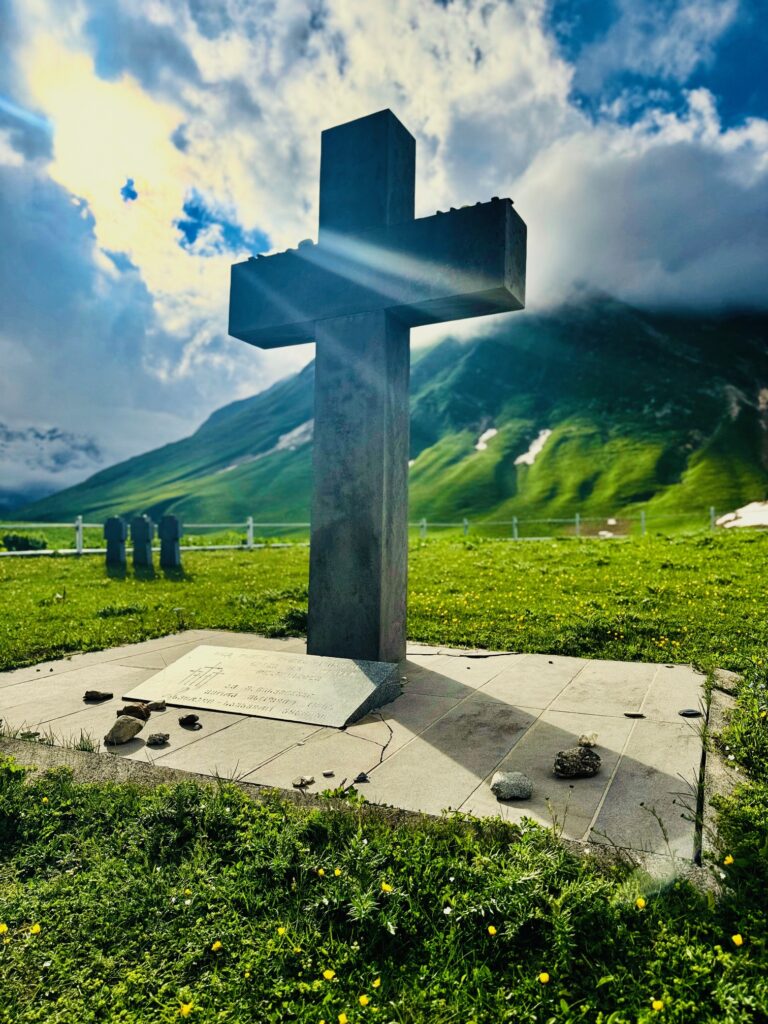Großes steinernes Kreuz auf einem gepflasterten Grab mit Berglandschaft und bewölktem Himmel im Hintergrund, daneben weitere Kreuze auf Gräbern
