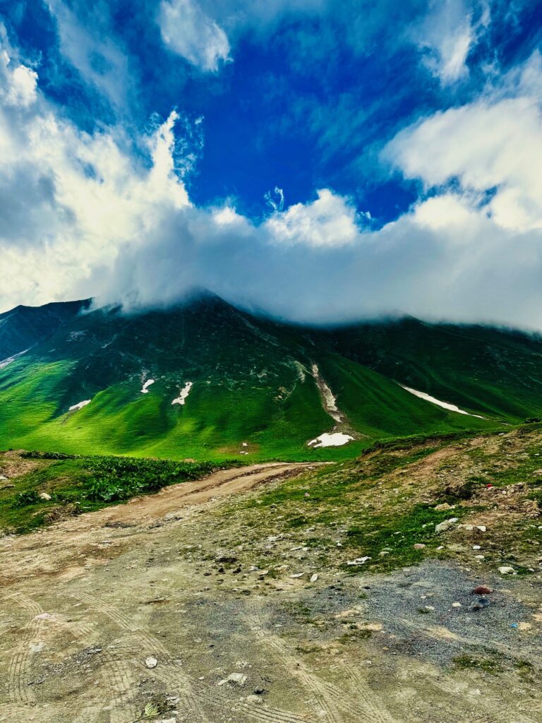 Weg vor einem grünen Berg, dessen Gipfel von Wolken bedeckt ist, unter blauem Himmel mit weißen Wolken