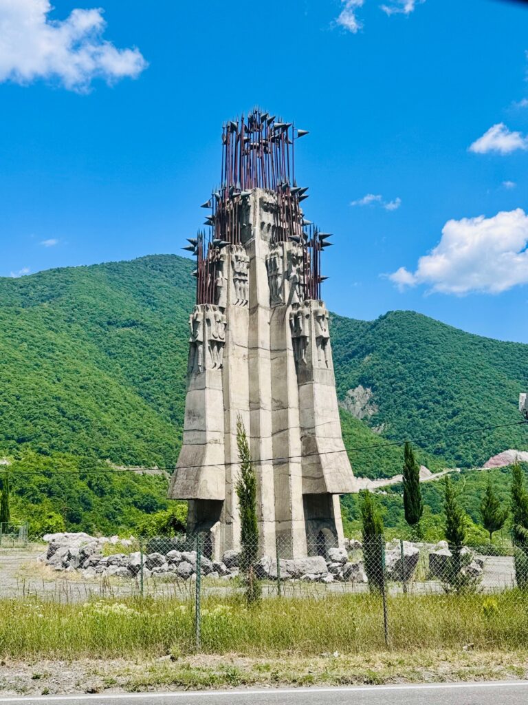Betonmonument mit stählernen Verstrebungen auf einer Wiese vor bewaldeten Bergen unter blauem Himmel