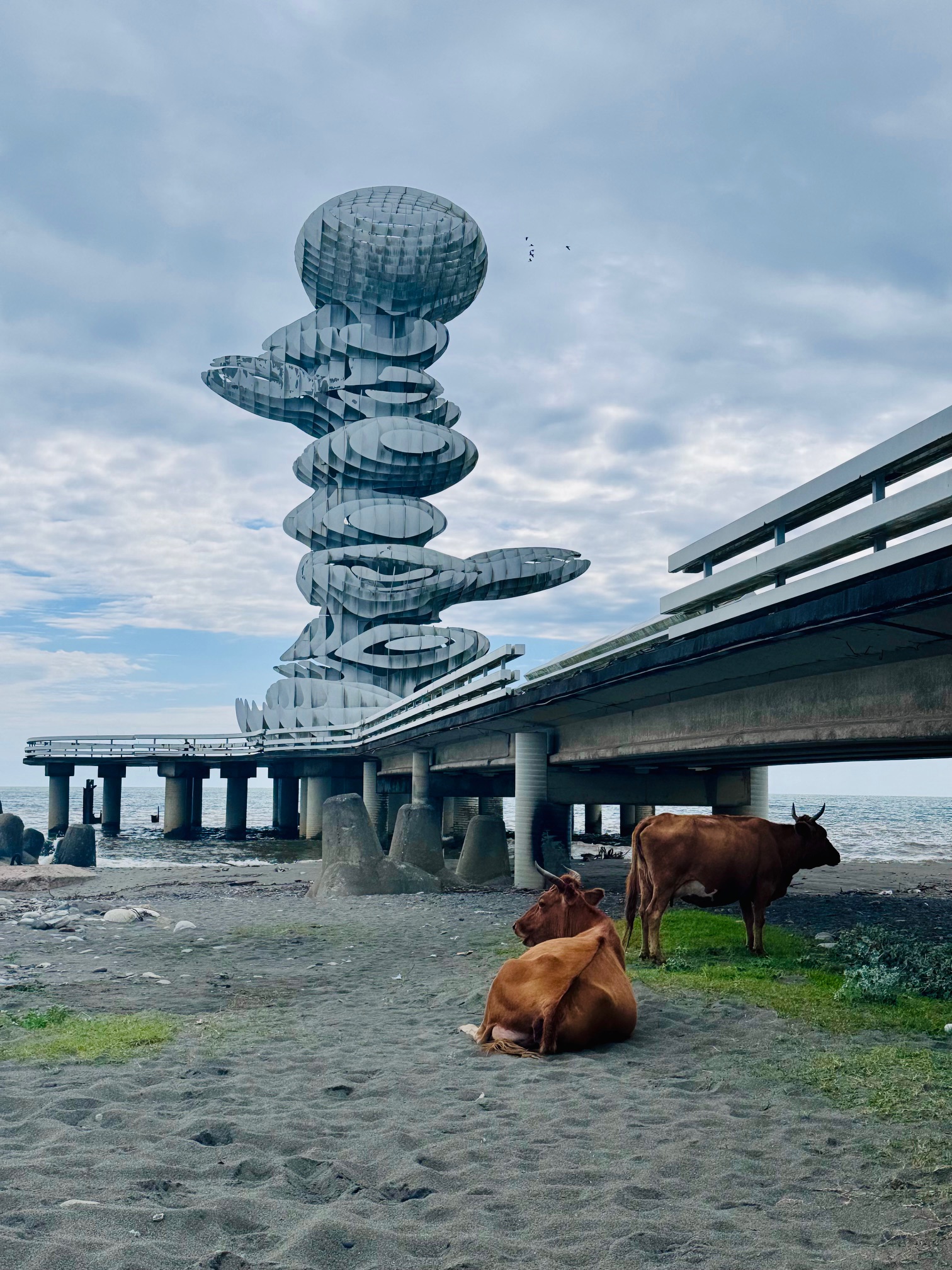 Skulptur aus mehreren übereinander angeordneten, ovalen, metallisch wirkenden Scheiben auf einem Steg am Strand mit zwei Kühen im Vordergrund