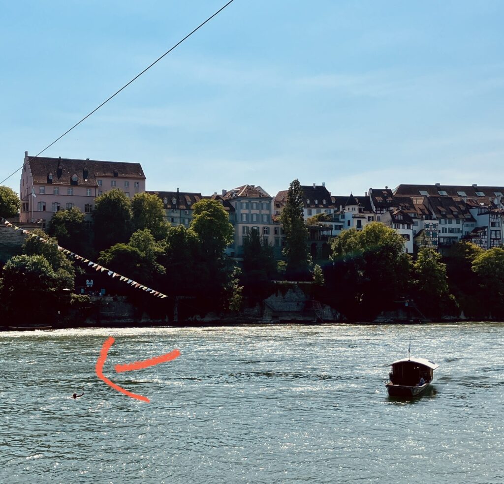 Der Rhein in Basel mit historischen Gebäuden im Hintergrund, Fähre auf dem Wasser und roter Pfeil, der auf eine Person im Wasser zeigt