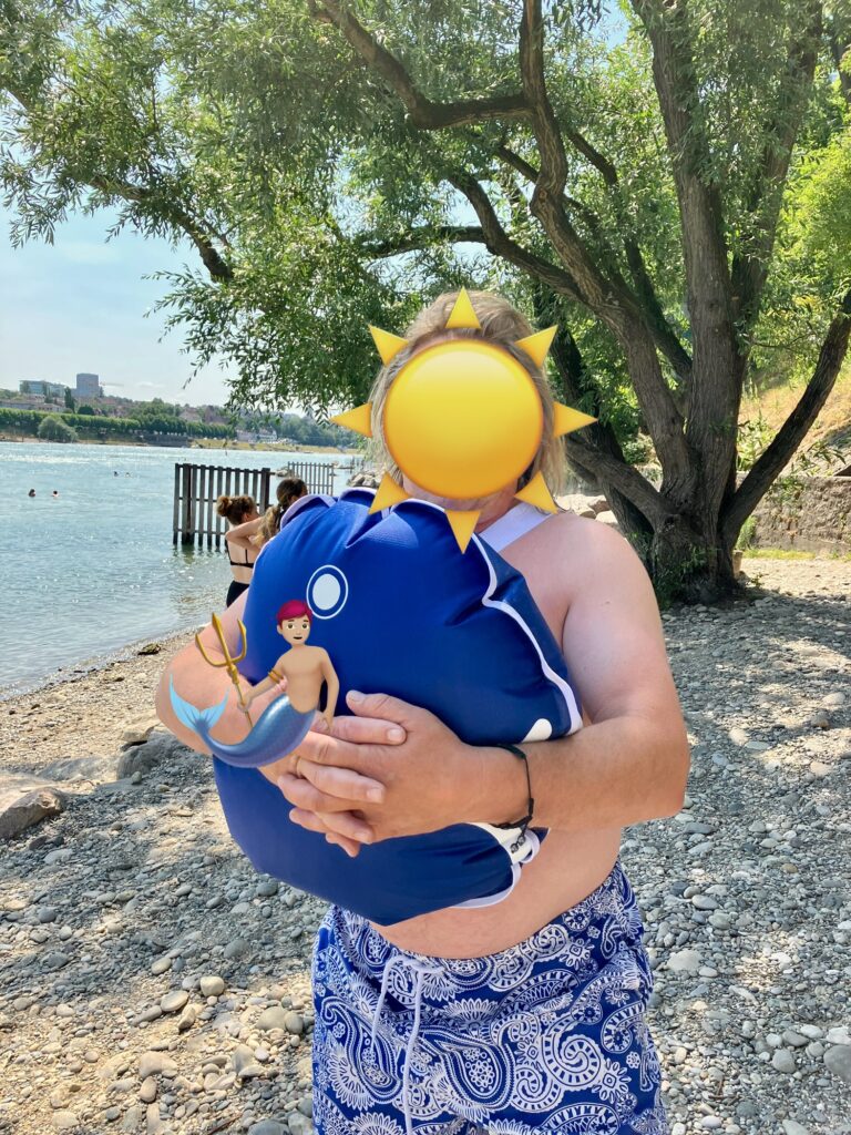 Person am Kiesstrand hält blauen Wickelfisch vor einem Baum und Fluss im Hintergrund