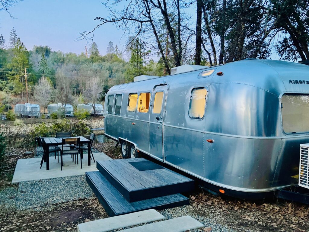 Silberner Airstream-Wohnwagen mit beleuchteten Fenstern steht auf einem Platz mit Holzstufen und einem Tisch mit Stühlen umgeben von Bäumen