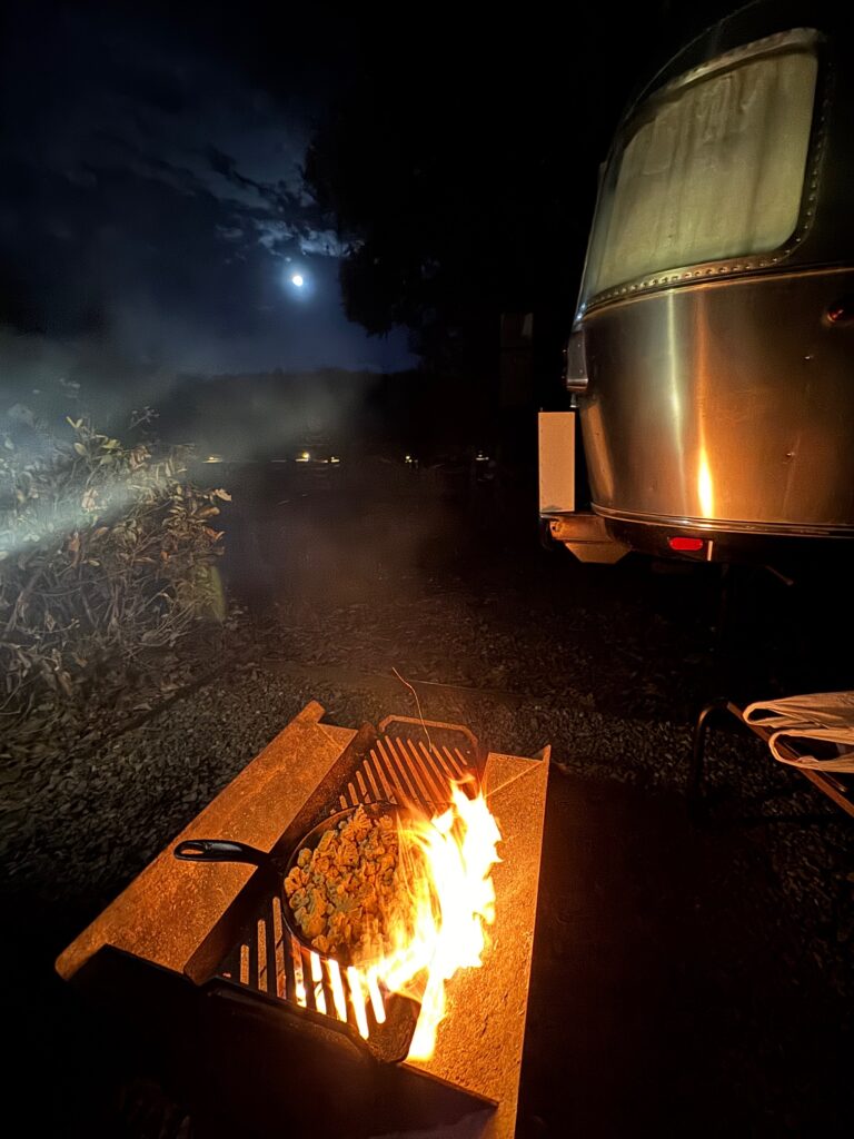 Lagerfeuer in einer Feuerstelle vor einem Airstream-Wohnwagen bei Nacht mit Vollmond und Wolken