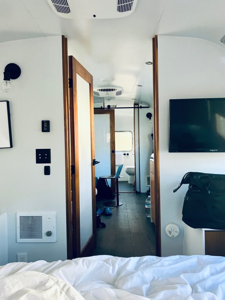 Innenraum eines modernisierten Airstream-Wohnwagens - Blick vom Bett durch einen schmalen Flur mit offenem Holztürrahmen in einen Raum mit Stuhl und Toilette