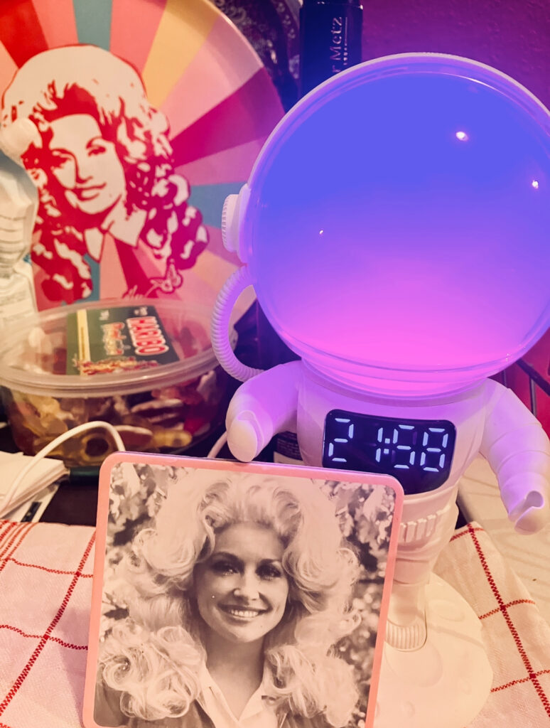 Astronauten-Wecker mit leuchtendem Helm und digitaler Uhrzeit 21:48 vor schwarz-weißem Porträt einer Frau mit voluminösem lockigem Haar auf einem Tisch mit kariertem Tuch, davor ein Foto von derselben Frau. Die Frau ist Dolly Parton
