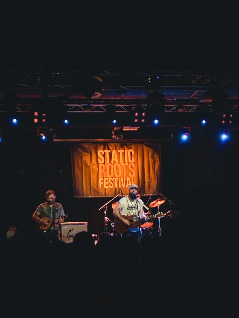 Zwei Musiker mit Gitarren auf einer Bühne vor einem Banner mit der Aufschrift 'STATIC ROOTS FESTIVAL'