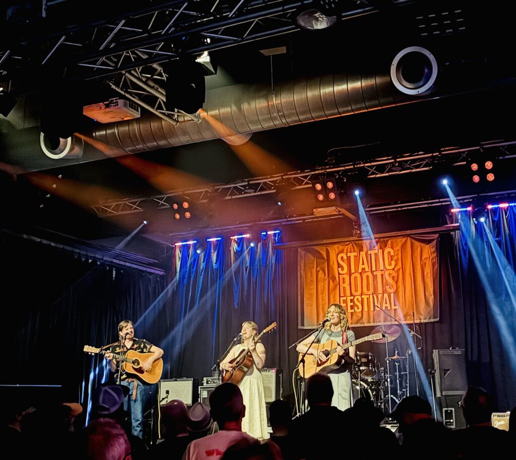 Drei Musiker mit Akustikgitarren auf einer Bühne vor einem Banner mit der Aufschrift 'STATIC ROOTS FESTIVAL' und Publikum im Vordergrund