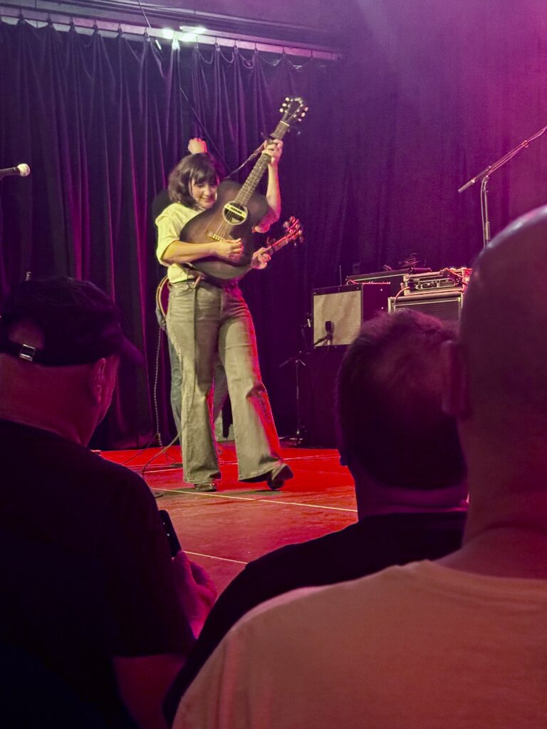 Musikerin mit Gitarre auf Bühne vor Publikum, roter Bühnenboden und schwarzer Vorhang im Hintergrund