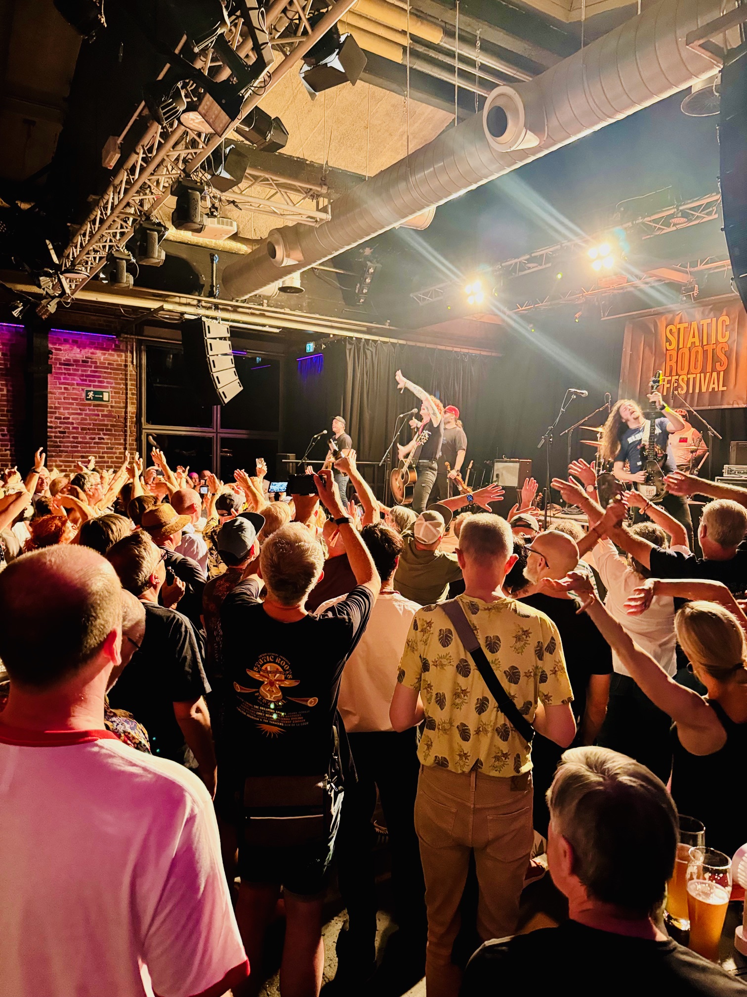 Publikum vor Bühne bei Konzert mit Band und heller Bühnenbeleuchtung in einer kleinen Fabrikhalle