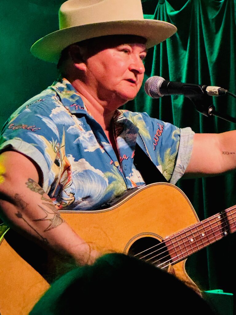 Person mit Hut und kurzärmligem Hemd spielt Gitarre und singt vor grünem Vorhang