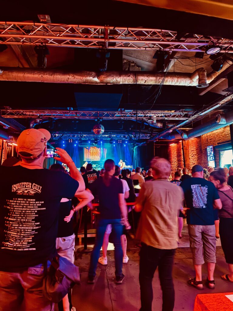 Publikum in einem Club mit Bühne und bunten Lichtern, einige Personen tragen T-Shirts mit Texten auf dem Rücken