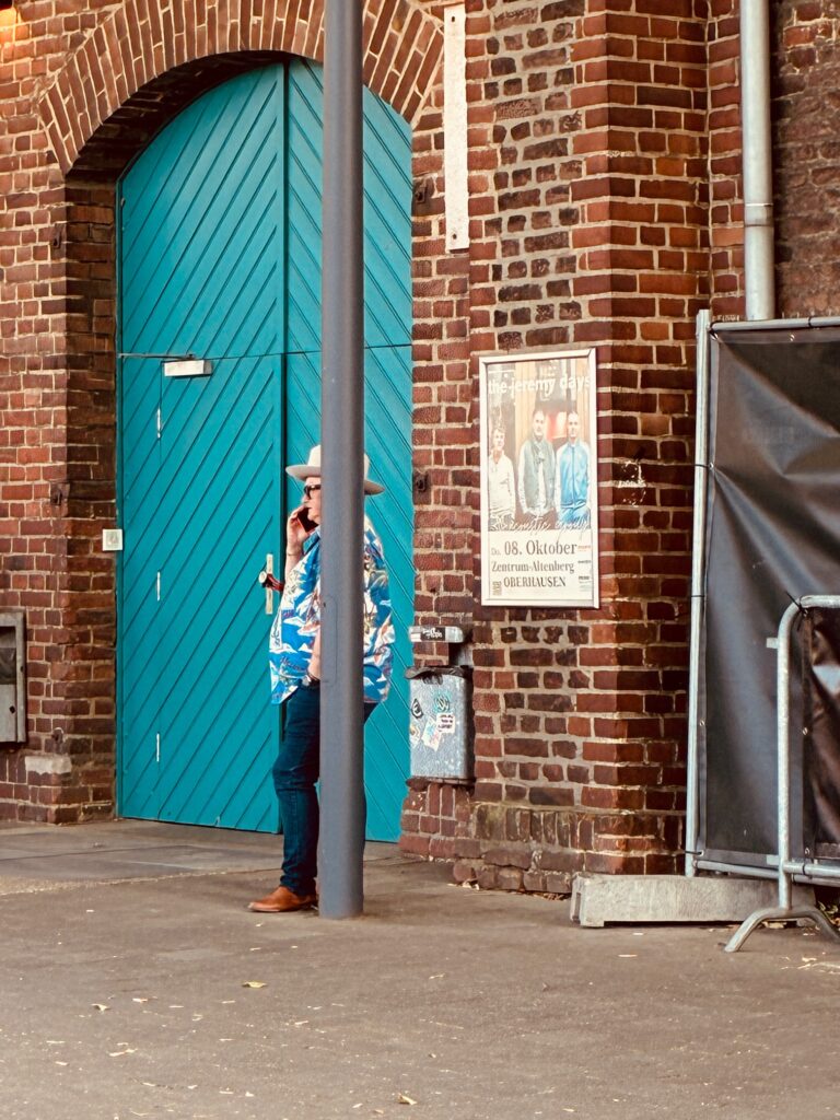 Person mit Hut und Hemd mit blau-weißem Muster lehnt an Laternenpfahl vor einer Backsteinwand mit blauem Holztor und Konzertplakat
