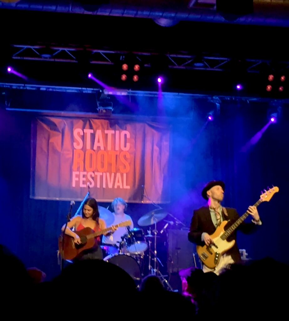 Drei Musiker auf einer Bühne beim Static Rocks Festival, eine Frau spielt Gitarre, ein Mann Bass, im Hintergrund ein Schlagzeuger