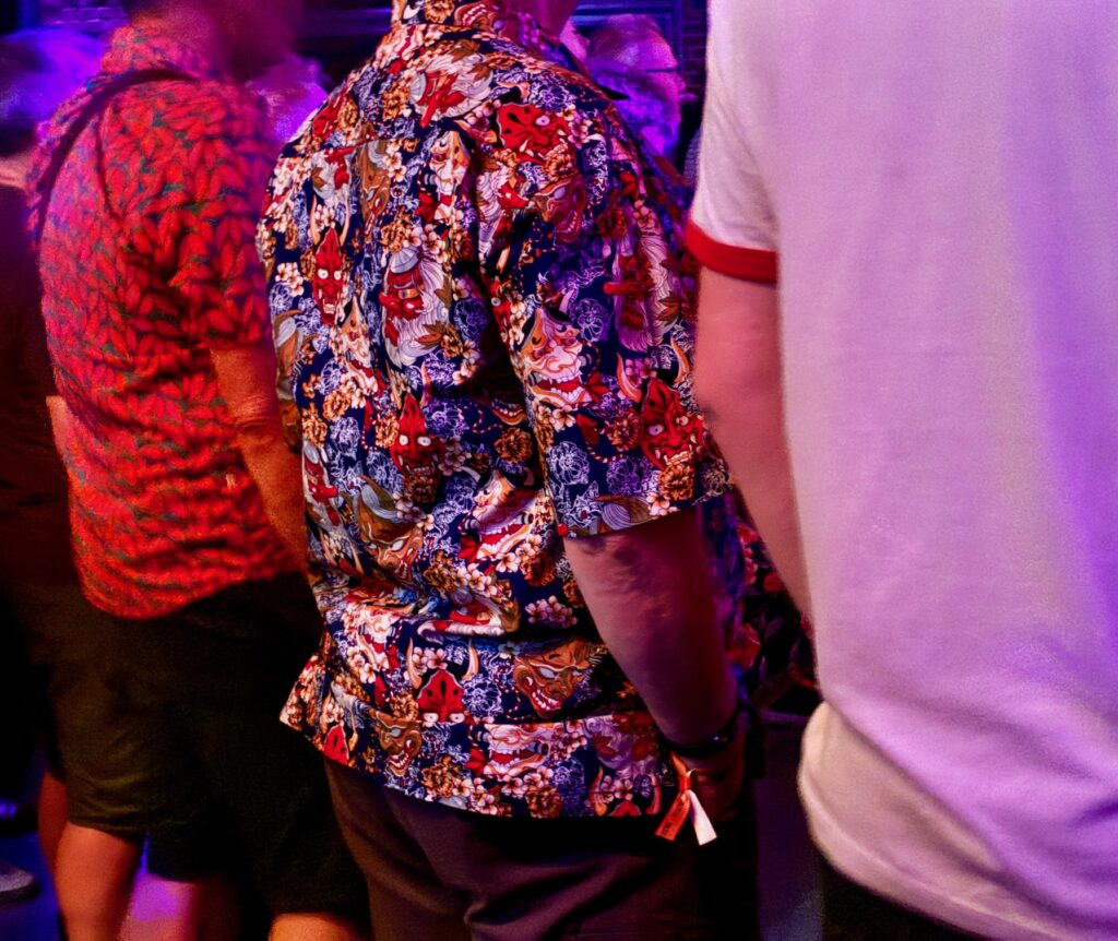 Drei Personen von hinten, eine trägt ein kurzärmeliges Hemd mit buntem Muster, eine ein weißes T-Shirt mit rotem Kragen und Ärmelabschluss, die dritte ein rot-schwarzes Hemd. Es sind nur Rücken und Arme zu sehen