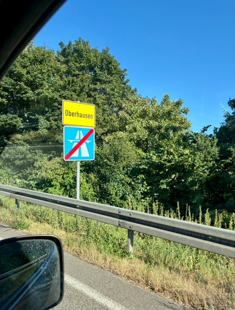 Gelbes Ortsschild mit der Aufschrift 'Oberhausen' über einem blauen Verkehrsschild, das eine durchgestrichene Autobahn symbolisiert, neben einer Straße mit Leitplanke und Bäumen im Hintergrund