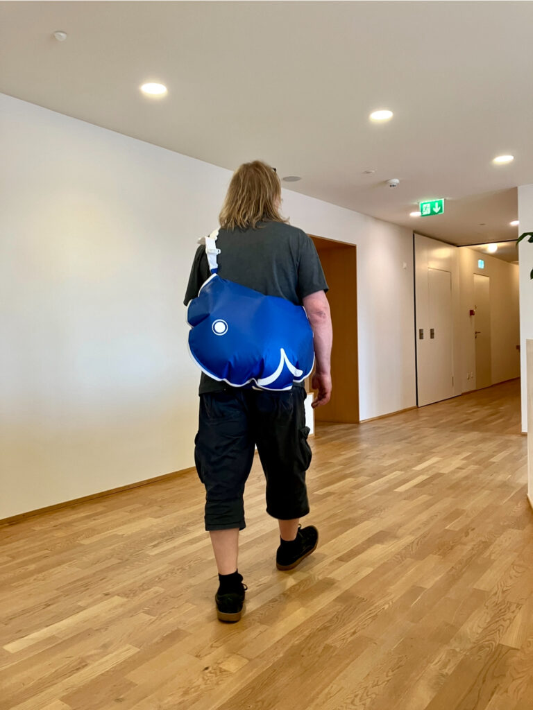 Person von hinten in einem Flur mit Holzfußboden trägt eine blaue, rundliche Tasche mit weißem Muster über der Schulter