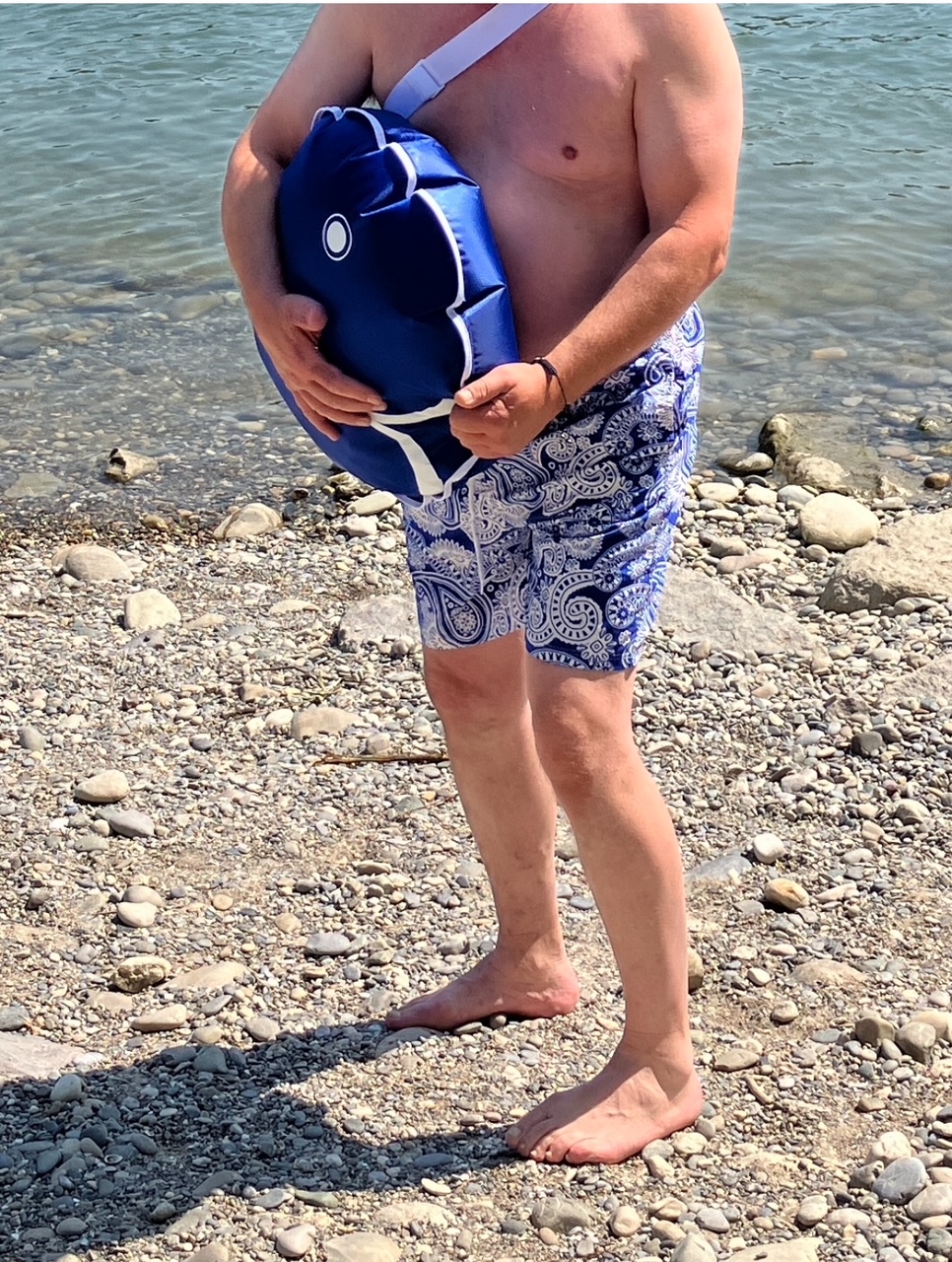 Oberkörperfreier Mann in gemusterten Badeshorts steht barfuß am kieseligen Ufer und hält eine blaue, gefüllte Schwimmtasche