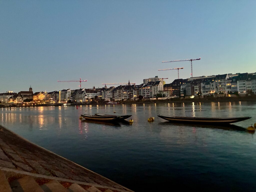 Uferpromenade mit mehreren Booten auf ruhigem Wasser und beleuchteten Gebäuden im Hintergrund bei Dämmerung