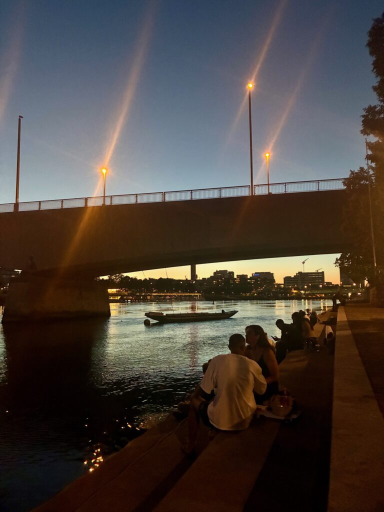 Menschen sitzen am Ufer des Rheins in Basel unter einer Brücke bei Dämmerung mit beleuchteten Straßenlaternen und einem Boot auf dem Wasser
