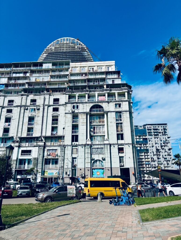Mehrstöckiges, marodes Wohngebäude mit Balkonen und zahlreichen Klimaanlagen davor parkende Autos und ein gelber Kleinbus bei blauem Himmel