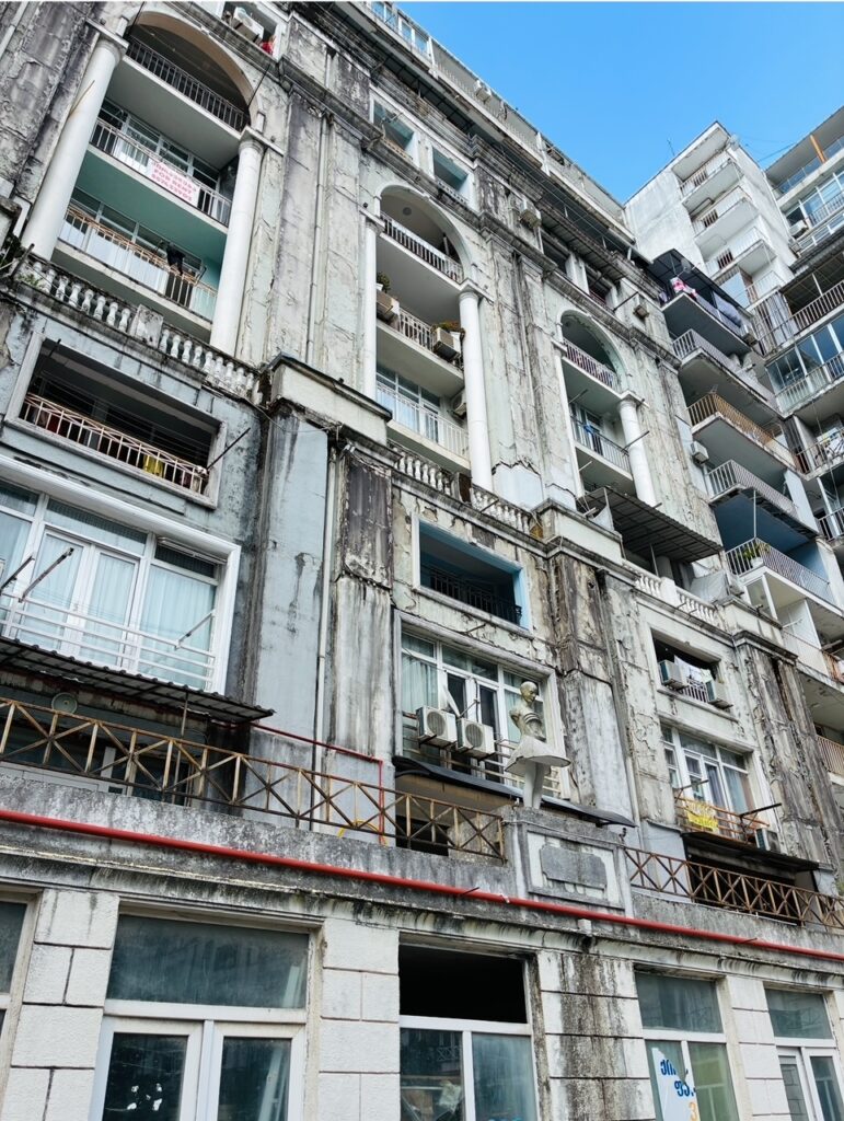 Verwitterte Fassade eines mehrstöckigen Gebäudes mit Säulen, Bogen, Balkonen, Fenstern und mehreren Klimageräten unter blauem Himmel