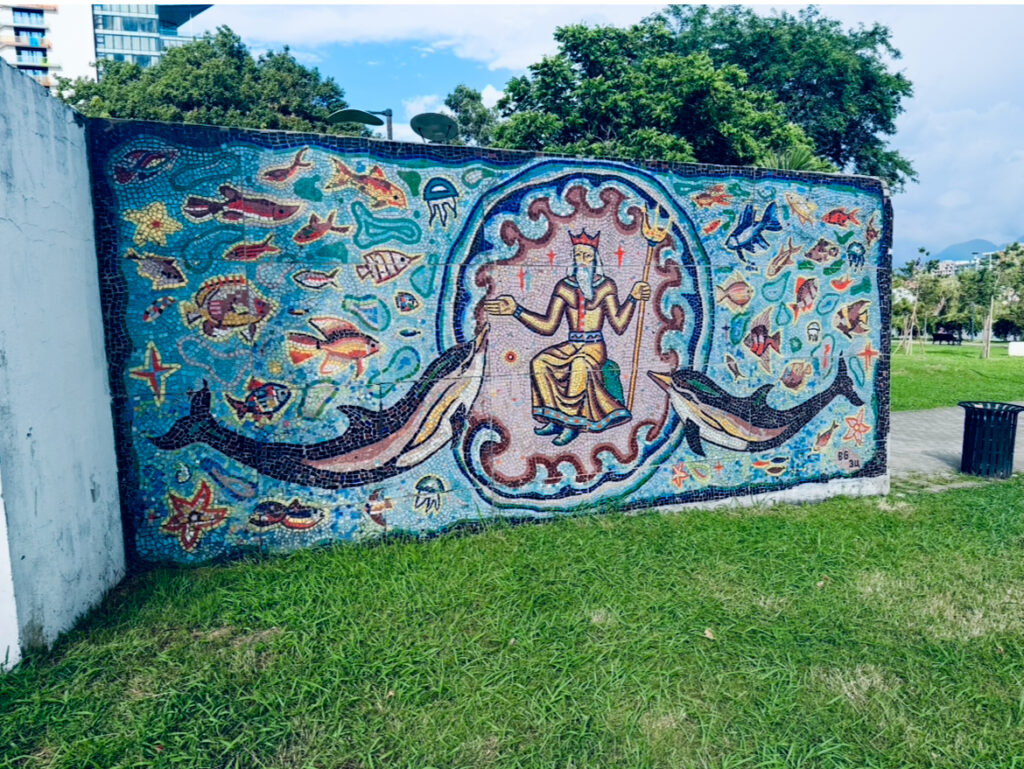 Mosaik an einer Außenwand zeigt eine zentrale Figur mit Krone und Dreizack, umgeben von Fischen, Delfinen und Meeresmotiven auf grünem Rasen vor blauem Himmel