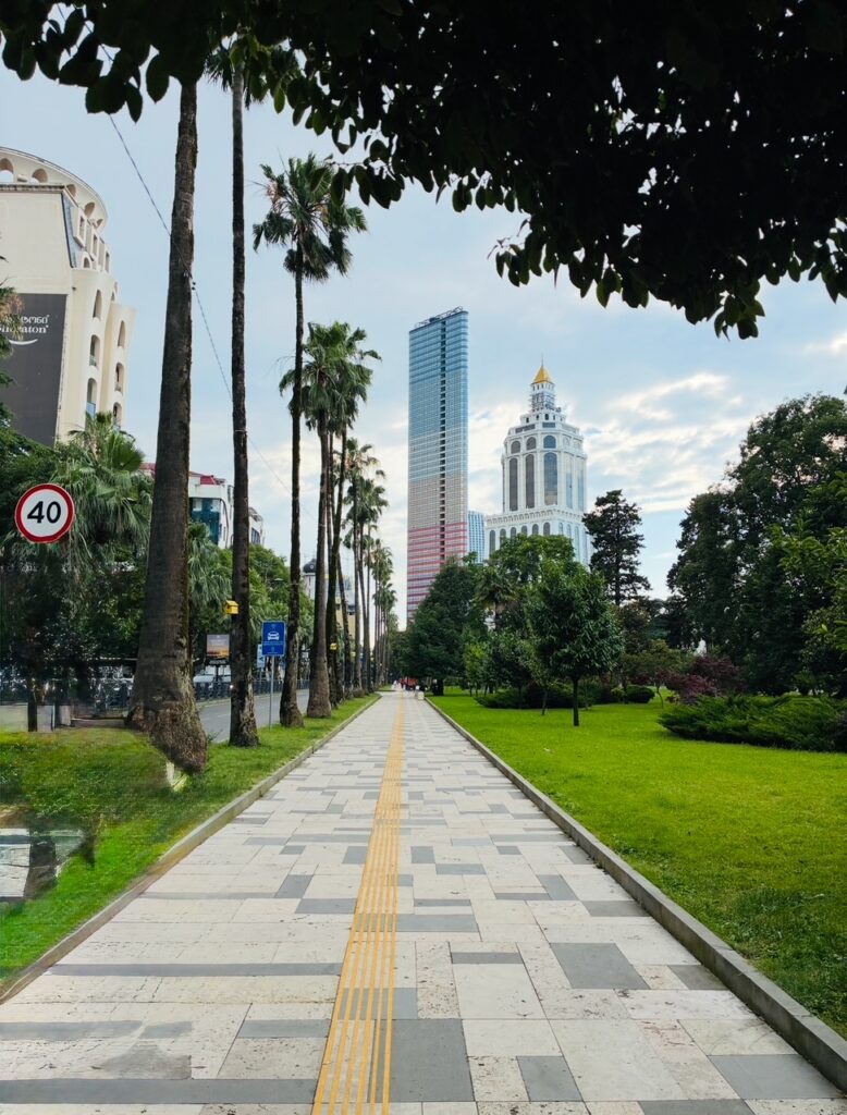 Breiter Fußweg mit gelber Leitlinie zwischen Palmen und Grünflächen vor modernen Hochhäusern unter blauem Himmel