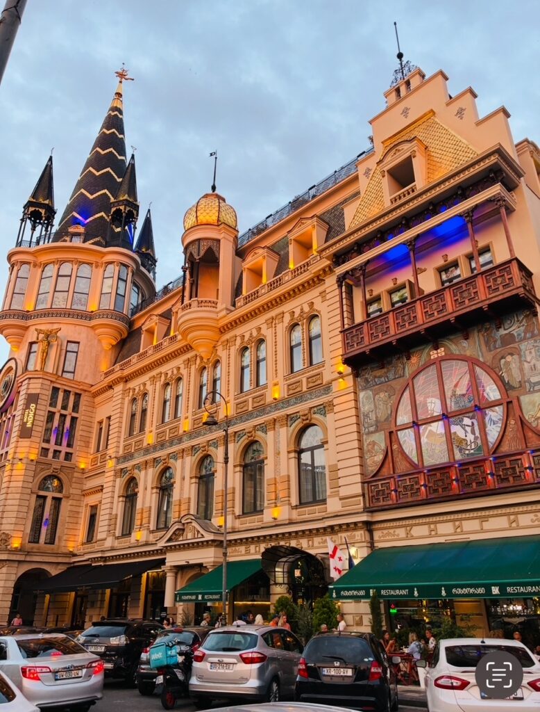 Historistisches Hotelgebäude mit Türmen, Erkern und beleuchteter Fassade, davor parkende Autos und Menschen an Straßencafés