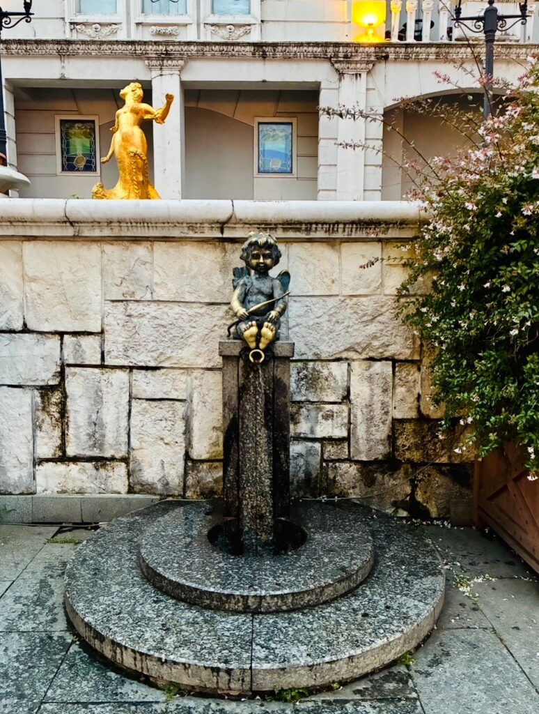 Brunnen mit einer bronzenen Kinderskulptur, die eine Trompete hält, vor einer steinernen Mauer mit einer goldfarbenen Statue und einem Busch rechts