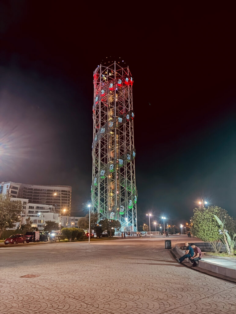 Hoher, beleuchteter Stahlgitterturm mit farbigen Lichtern in einer nächtlichen Stadtlandschaft mit Sitzbank und zwei Personen im Vordergrund
