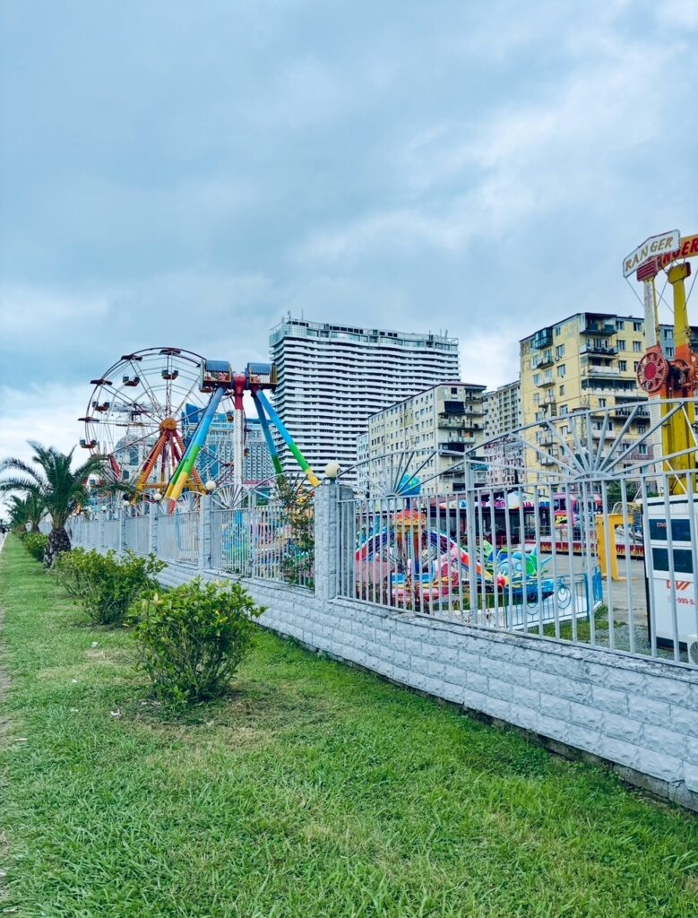 Kleiner Freizeitpark mit Riesenrad, Karussell und weiteren Fahrgeschäften hinter einem weißen Zaun vor Hochhäusern unter bewölktem Himmel