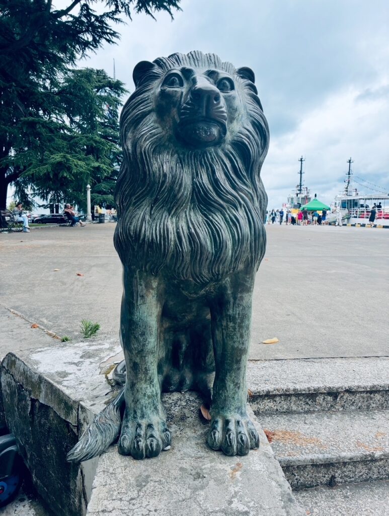 Bronzefigur eines sitzenden Löwen auf einem Steinpodest an einem Hafen mit Bäumen und Menschen im Hintergrund