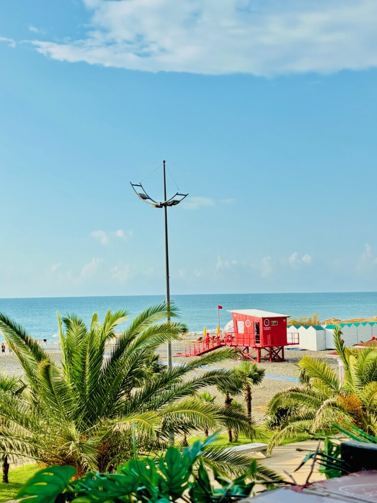 Strand mit Palmen im Vordergrund, rotem Rettungsturm und blauem Himmel über ruhigem Meer
