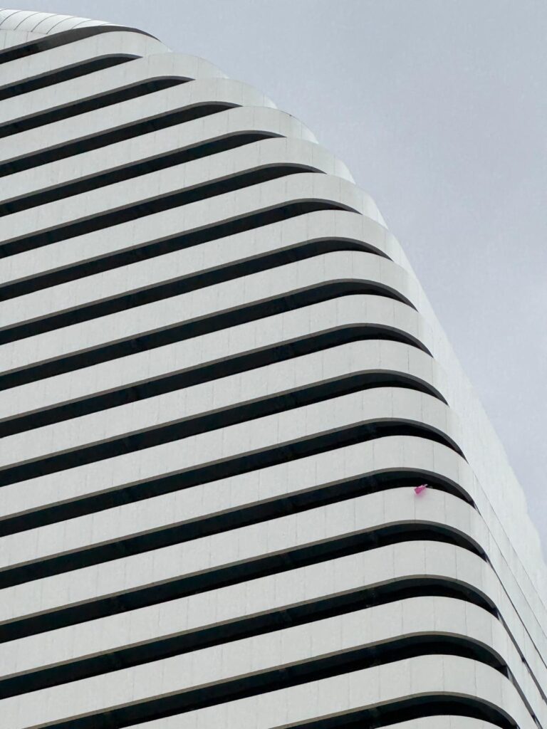 Fassade eines modernen Hochhauses mit geschwungenen horizontalen Linien und abgerundeten Ecken. Eine Etage ist mit einer kleinen rosa Fahne markiert