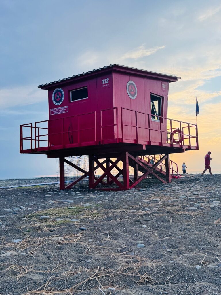 Rotes, erhöhtes Wachhaus auf Stelzen am steinigen Strand mit zwei Personen im Hintergrund