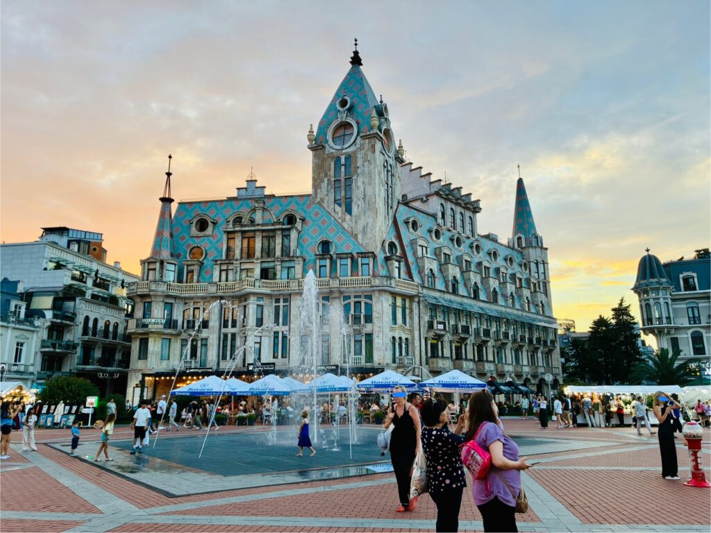 Historisch wirkendes Gebäude mit spitzem Turm und bunter Dachverkleidung an einem belebten Platz mit Springbrunnen und Menschen bei Sonnenuntergang