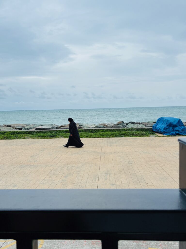 Eine in schwarzer Kleidung gehüllte Person geht auf einem gepflasterten Weg am Meer entlang, im Hintergrund Wolkenhimmel und Felsen am Ufer