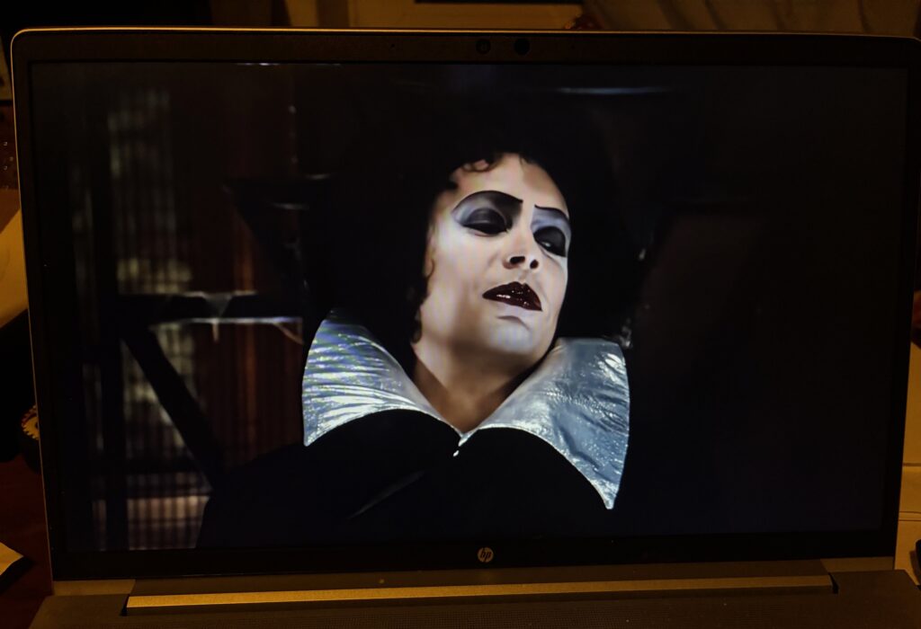 Nahaufnahme einer Person mit dunklem Lippenstift, stark geschminkten Augen und einem silbernen Kragen vor dunklem Hintergrund