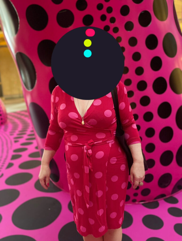 Frau in rotem Kleid mit rosa Punkten vor einer Wand mit großem pink-schwarzem Punktemuster. Kopf der Frau ist mit schwarzem Punkt verdeckt